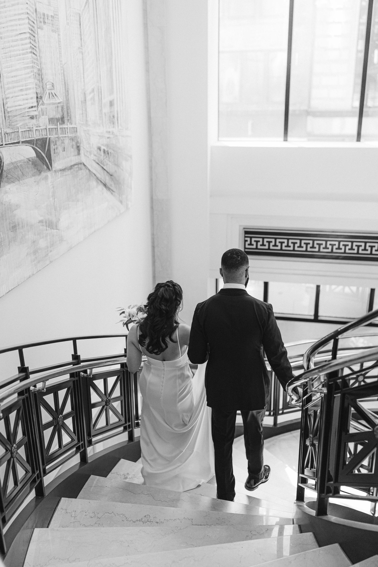 A bride and groom descend an elegant staircase with ornate railings in black and white.