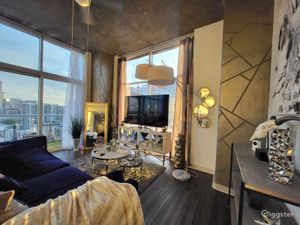 Elegant loft living room with gold accents, navy velvet sofa, floor-to-ceiling windows, and stunning city views at dusk.