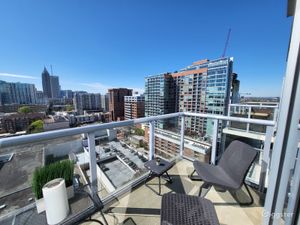 Sunny rooftop balcony with seating chairs overlooking a vibrant urban cityscape with blue skies and modern buildings.