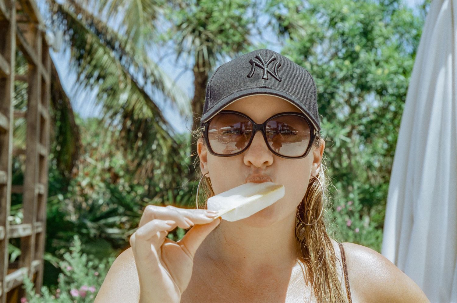 A person in sunglasses and baseball cap enjoying a popsicle in a tropical setting.