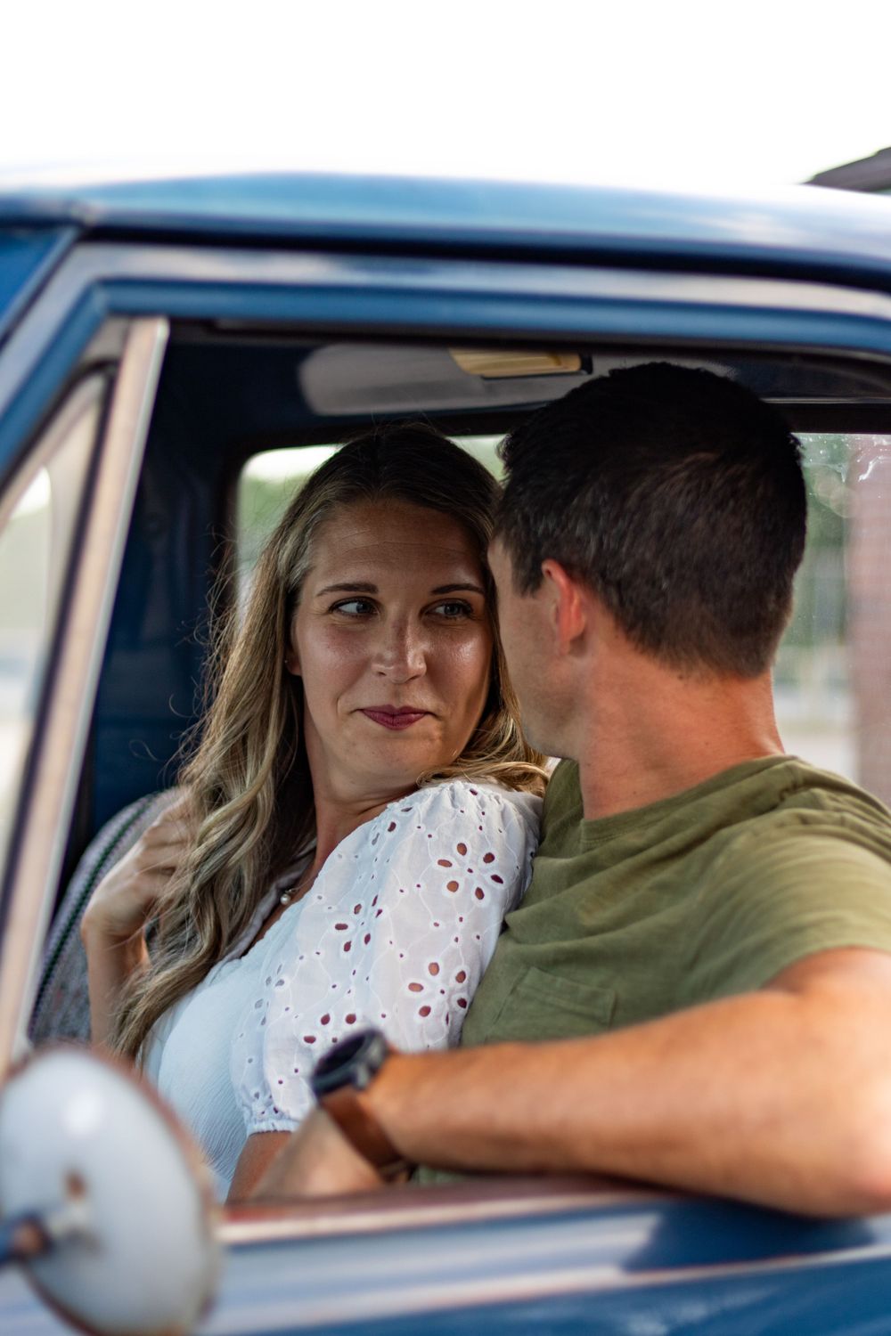 Couple sitting together in vintage car with white eyelet dress visible.