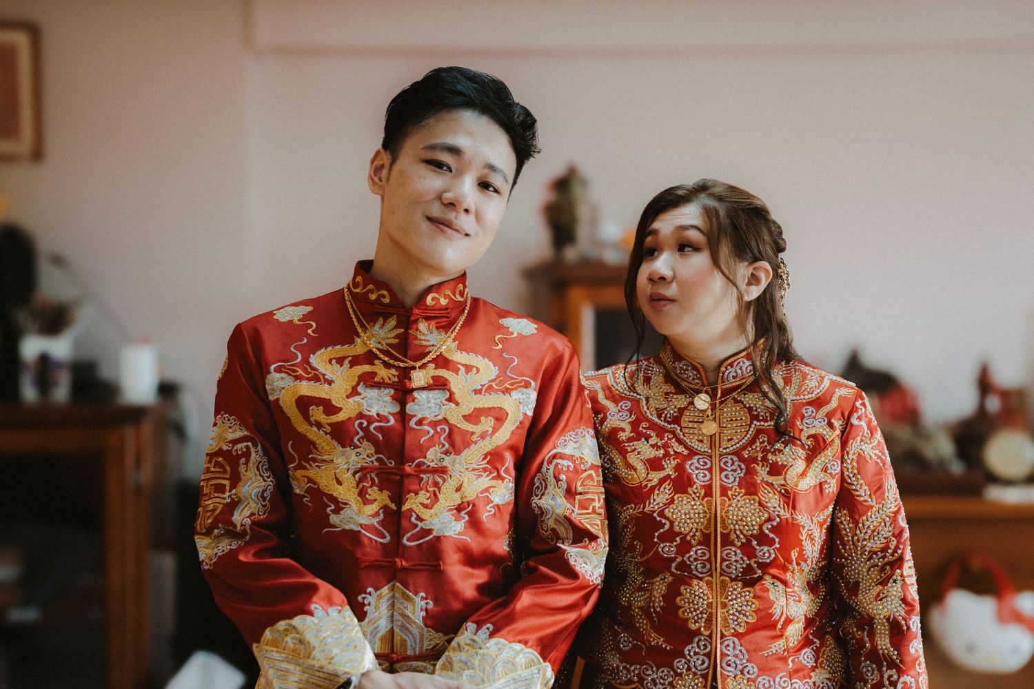 Couple wearing traditional red Chinese wedding attire with gold embroidery in a home setting.