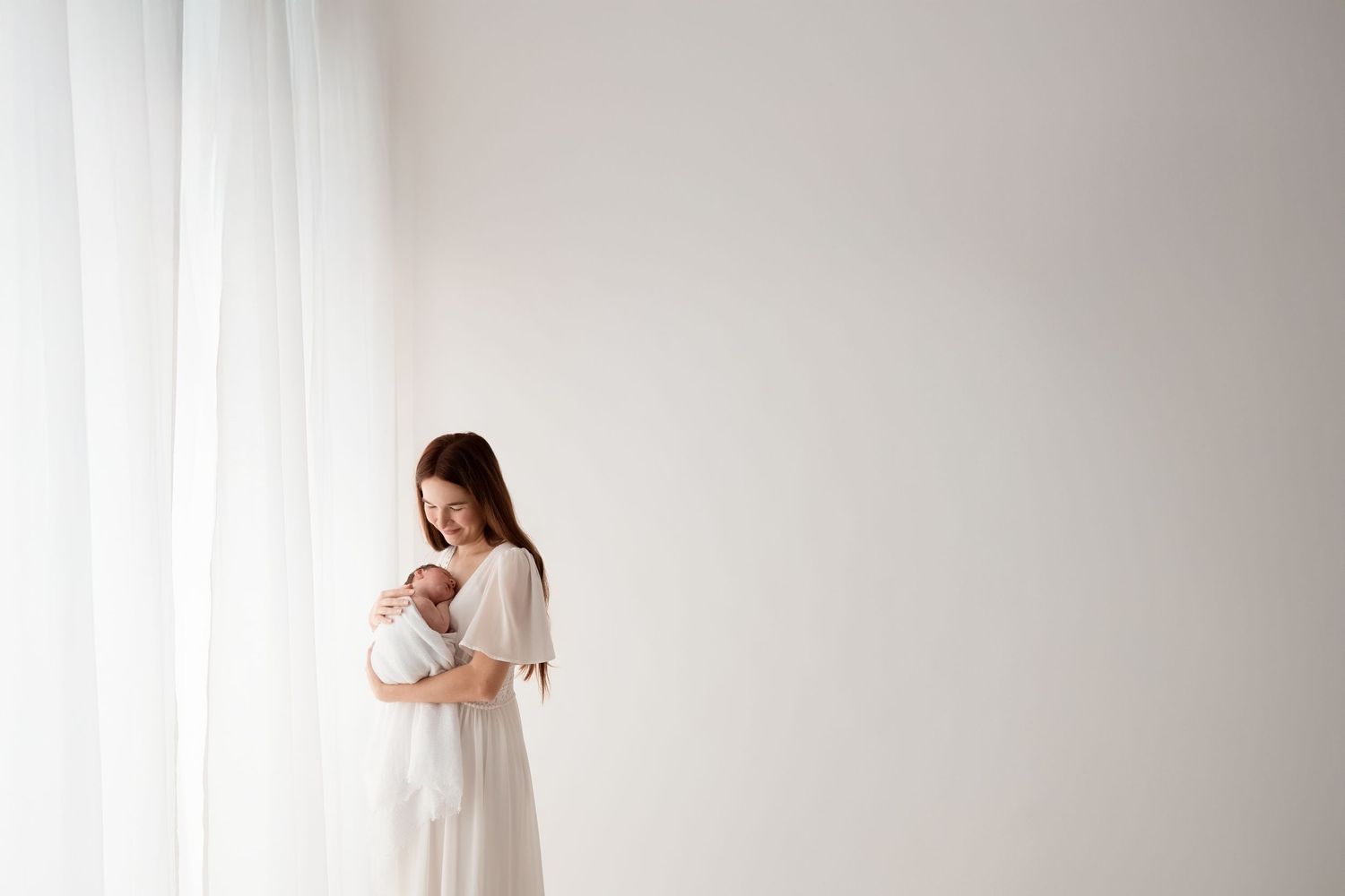 Young mother in white dress cradling her newborn baby near a bright window.