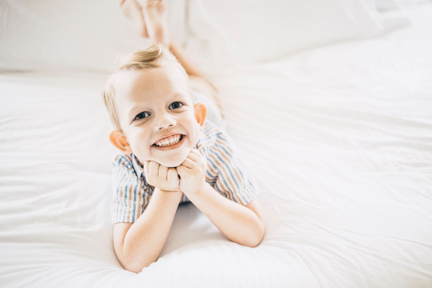 Happy smiling toddler boy lying on white bed with chin resting on hands.