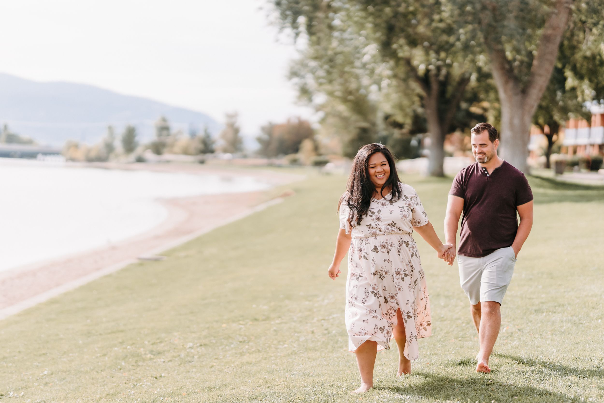 A couple strolls hand in hand along a grassy lakeside path on a sunny summer day.
