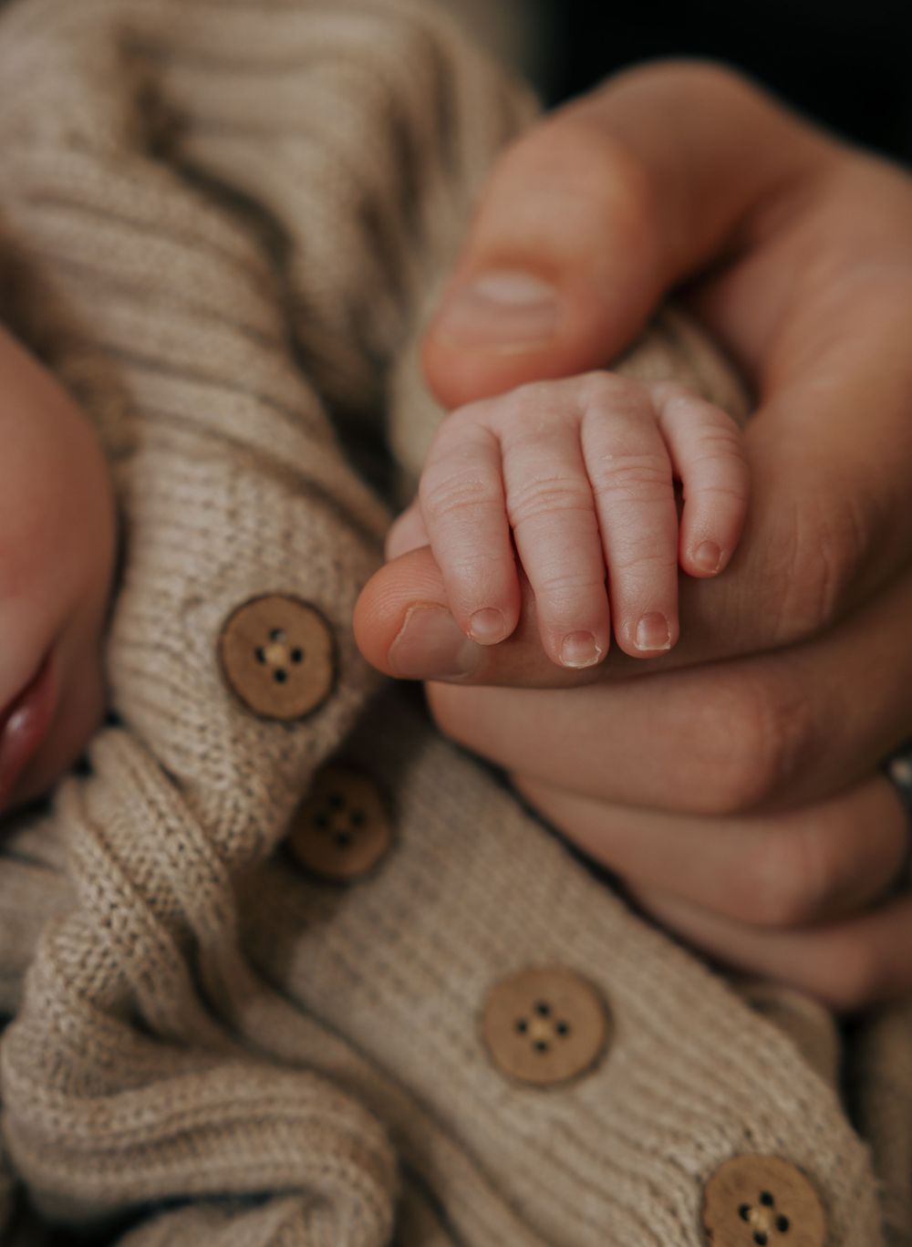Delicate newborn fingers hold onto adult hand beside beige knit sweater with buttons.