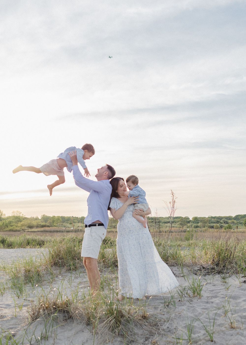 A loving family poses together at dusk on a sandy beach with grassy dunes and cloudy skies in the background.