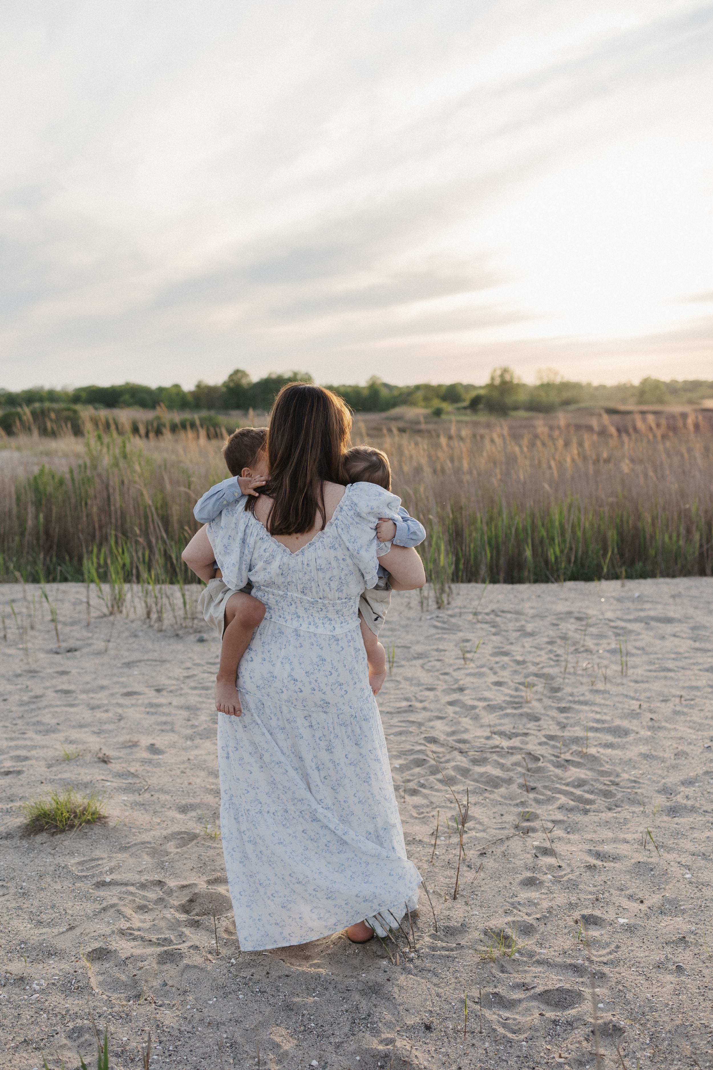 A person in a white floral dress sits on a sandy beach at sunset with tall beach grass in the background.