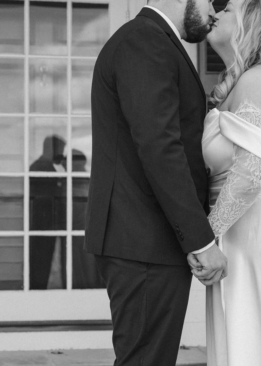A couple in wedding attire shares an intimate moment in front of a window during their black and white wedding photoshoot.
