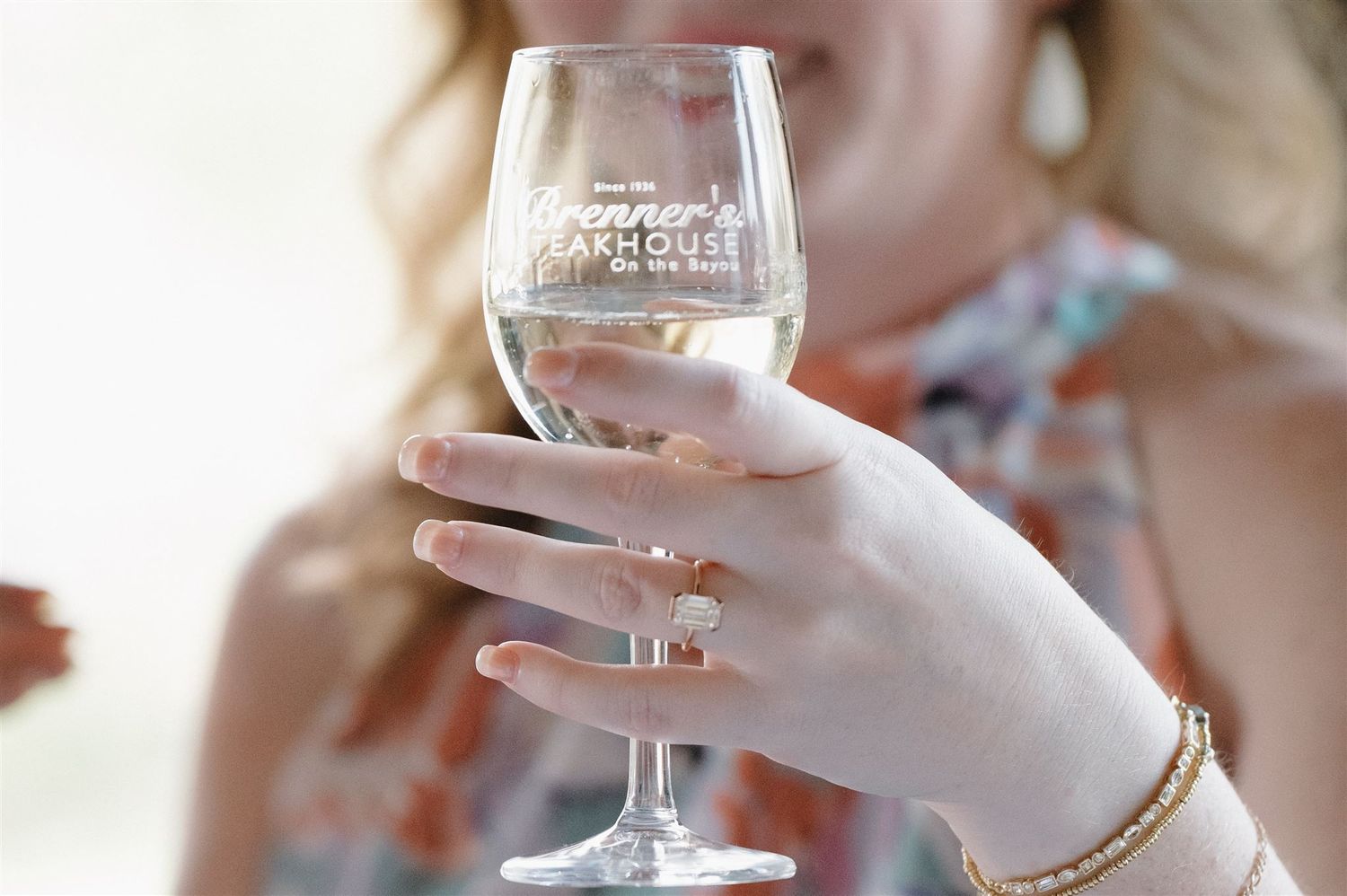 A manicured hand wearing gold jewelry holds a wine glass in natural light.