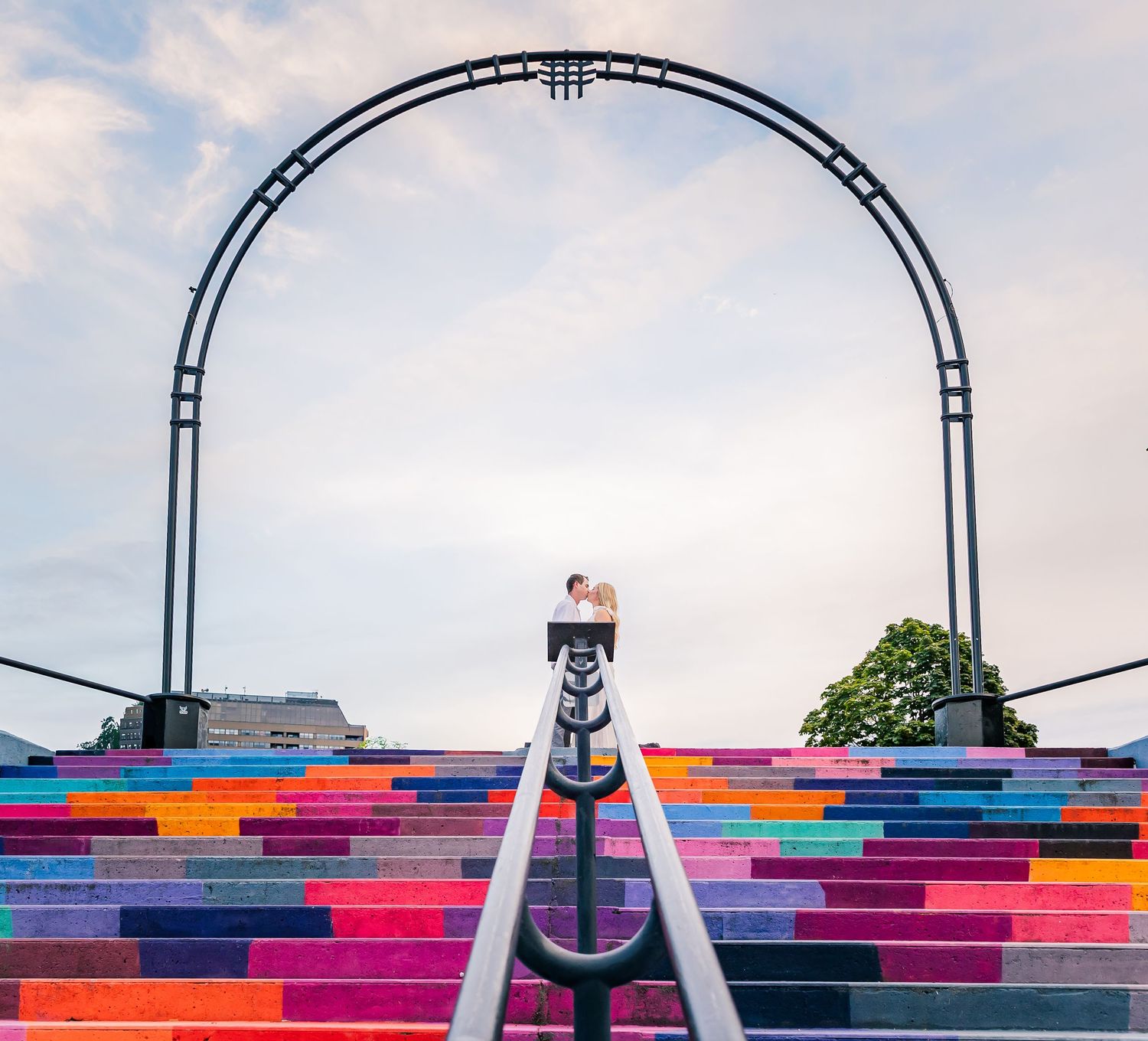 Colorful rainbow stairs lead up to a black metal arch against a cloudy sky.