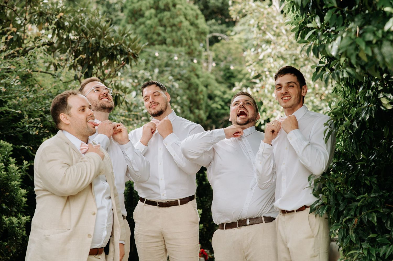 Groomsmen in light colored suits laughing and adjusting their bowties.