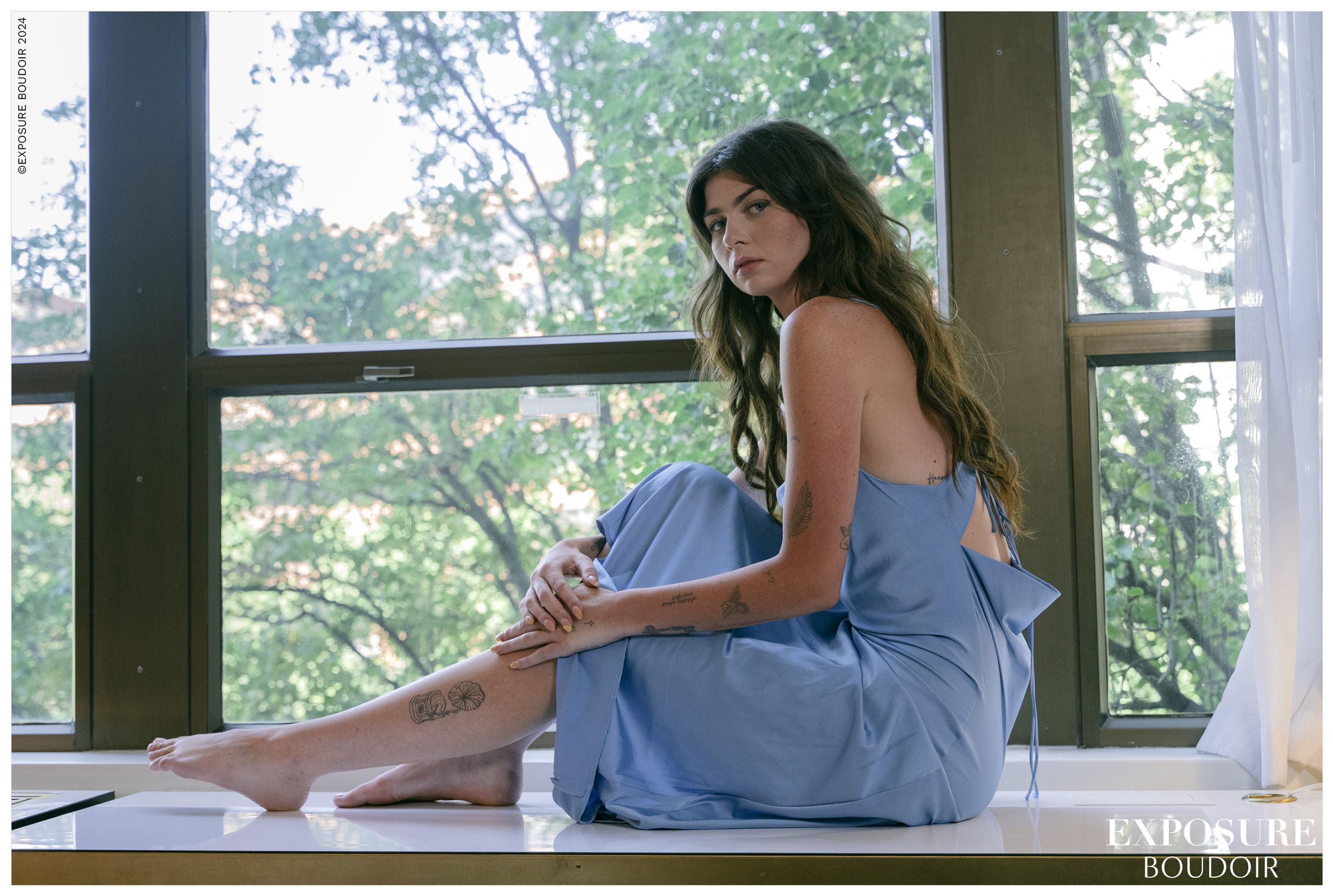 Someone in a light blue dress sits thoughtfully on a wooden ledge by large windows in a sunlit room.