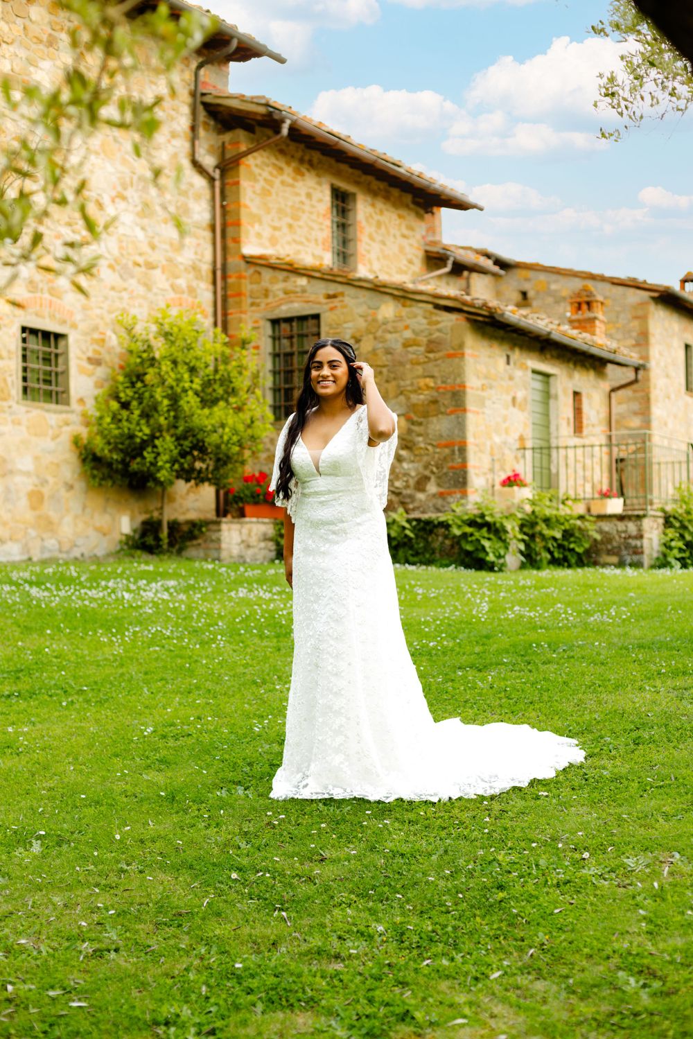 Bride in elegant white dress poses in front of rustic Tuscan villa with green lawn.