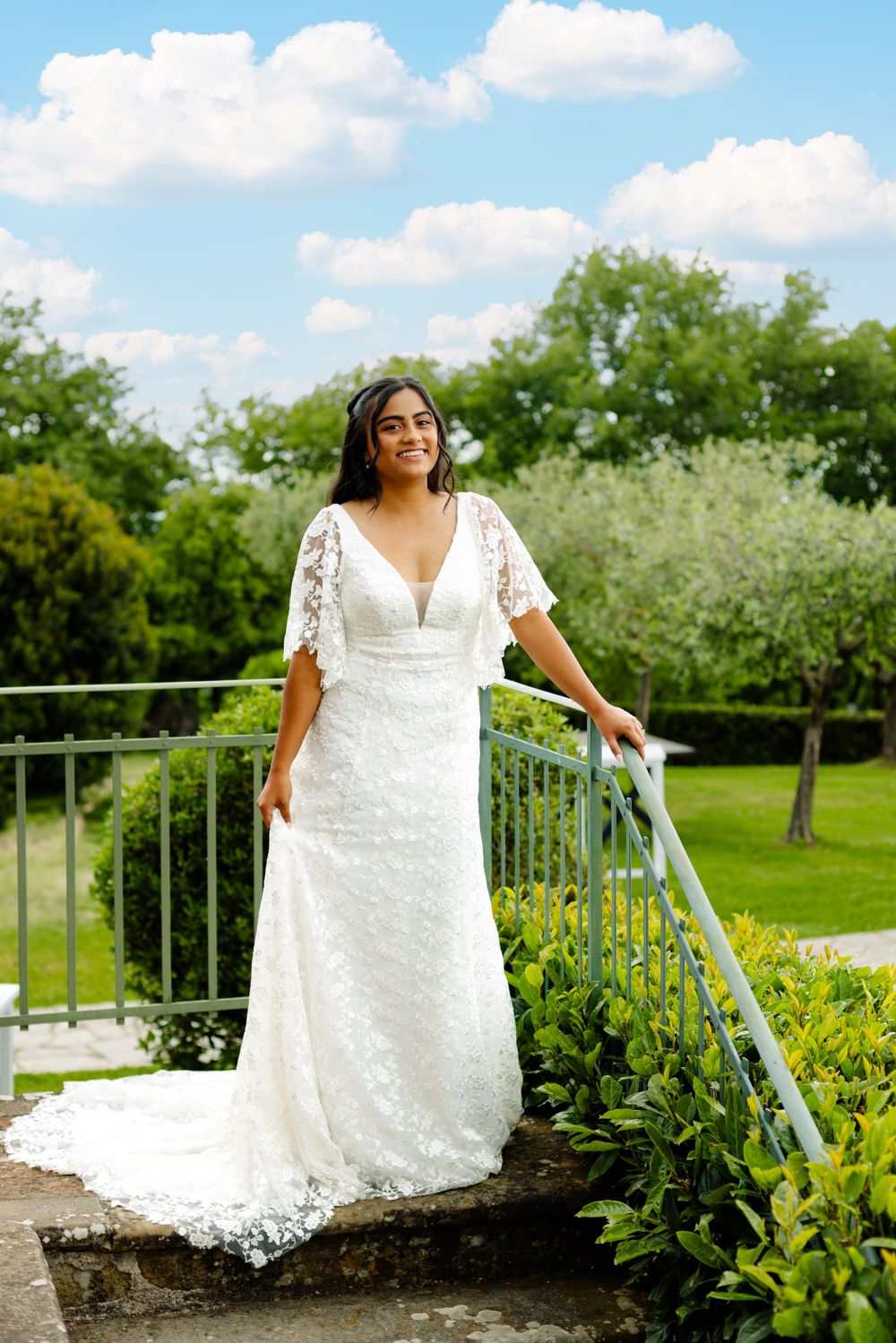 Bride in lace wedding dress poses elegantly on outdoor stone steps with garden backdrop.