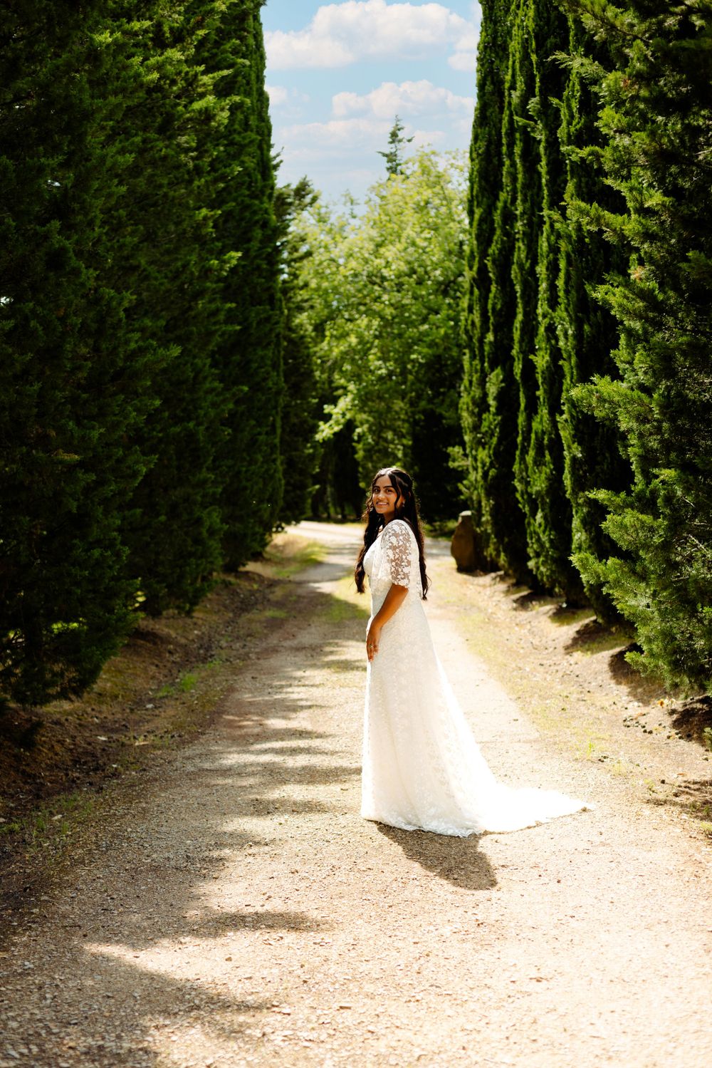 Bride in white wedding gown walks along cypress-lined path on sunny day.