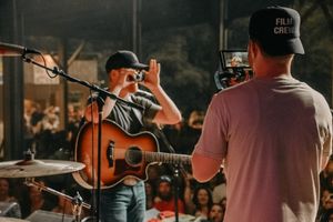A musician performs with an acoustic guitar on stage while being filmed by someone in a baseball cap.