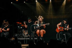 Musicians perform on stage with guitars and drums under dramatic concert lighting.
