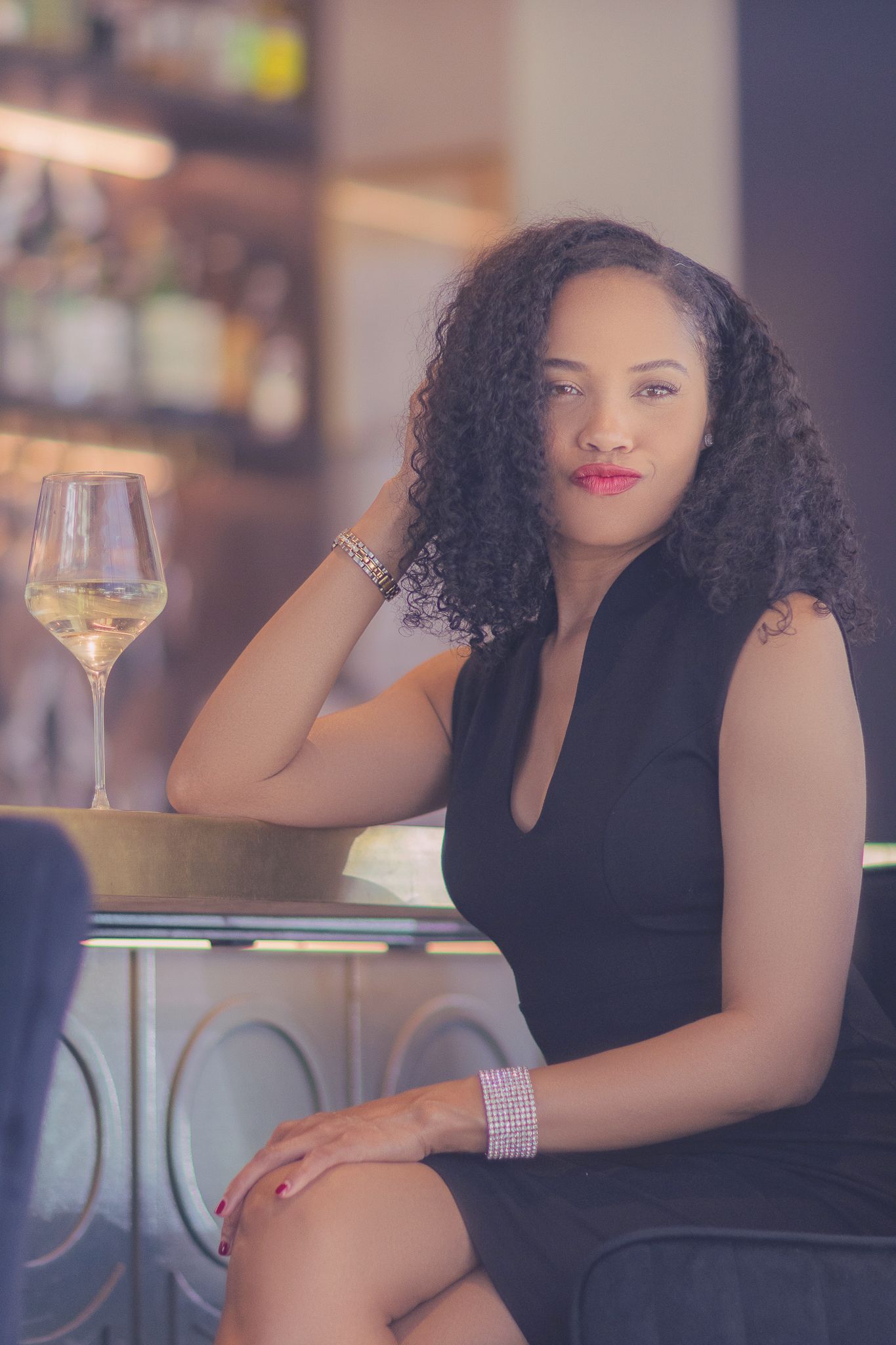 Elegant woman in black dress poses with glass of white wine at upscale restaurant bar.