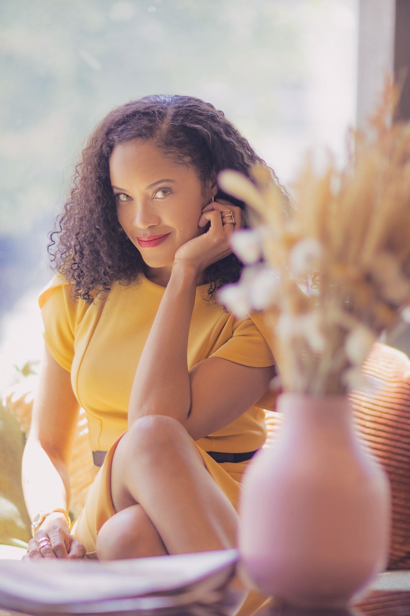 Person in yellow dress sits at table with vase of dried flowers in soft dreamy lighting.