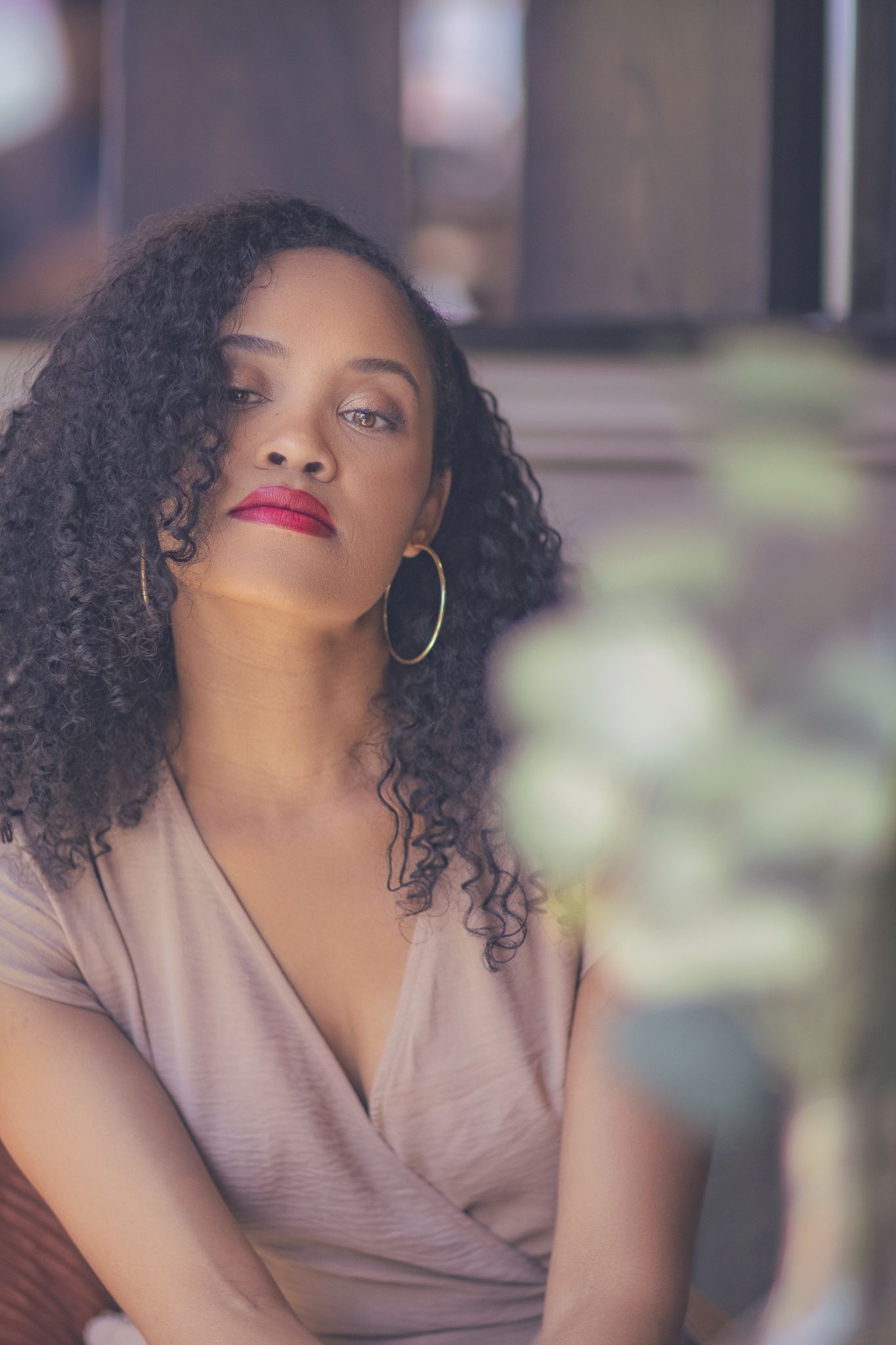 Portrait of someone with curly hair and gold hoop earrings wearing mauve wrap top gazing upward in soft lighting.