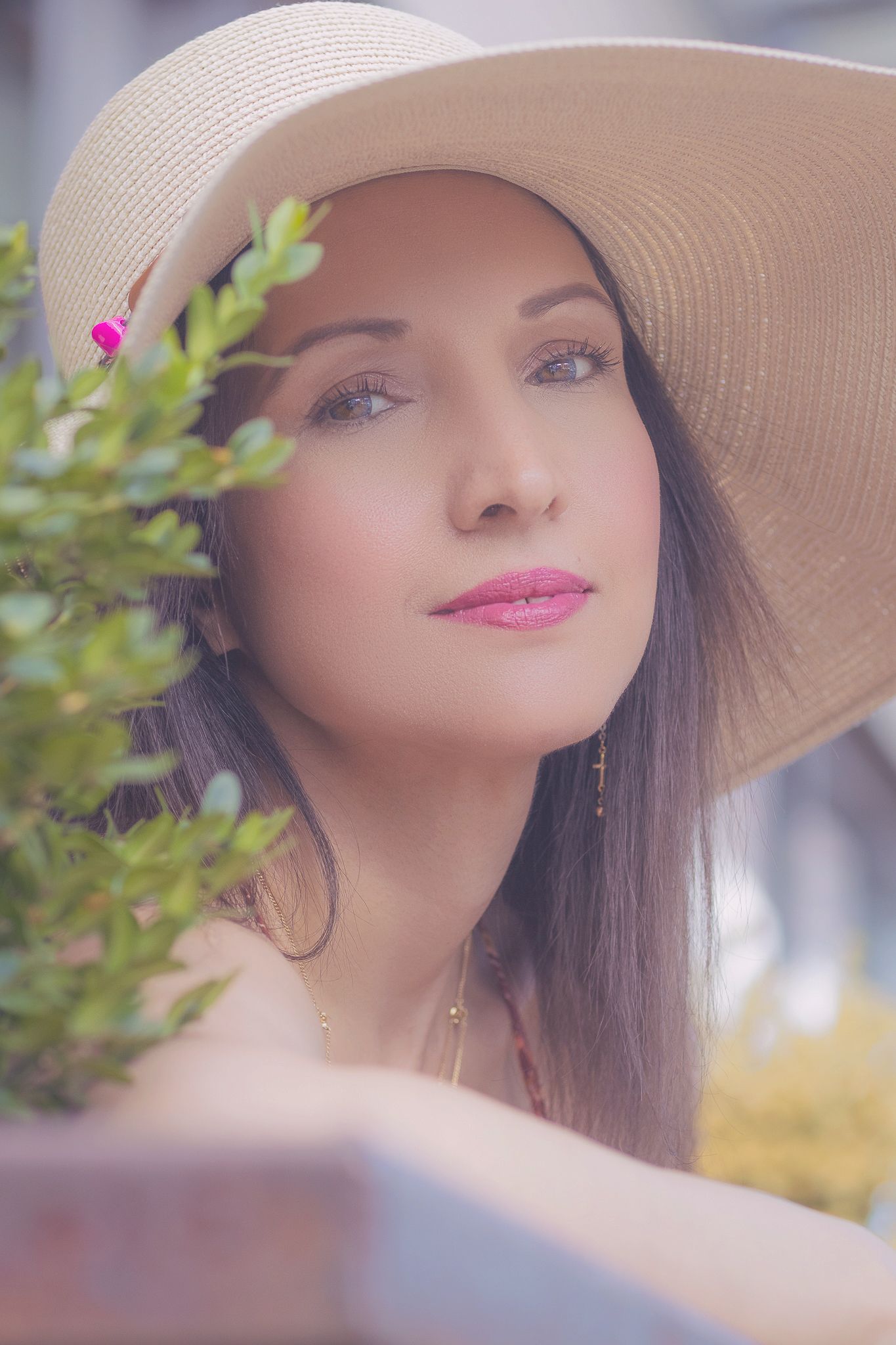 A dreamy summer portrait featuring a wide-brimmed beige sunhat, pink lipstick, and green foliage in soft focus.