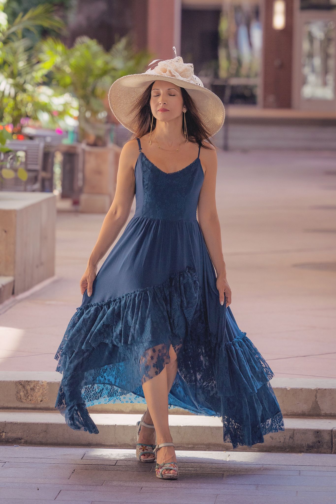 A woman in a flowing navy blue summer dress and beige sunhat walks along a plaza on a sunny day.