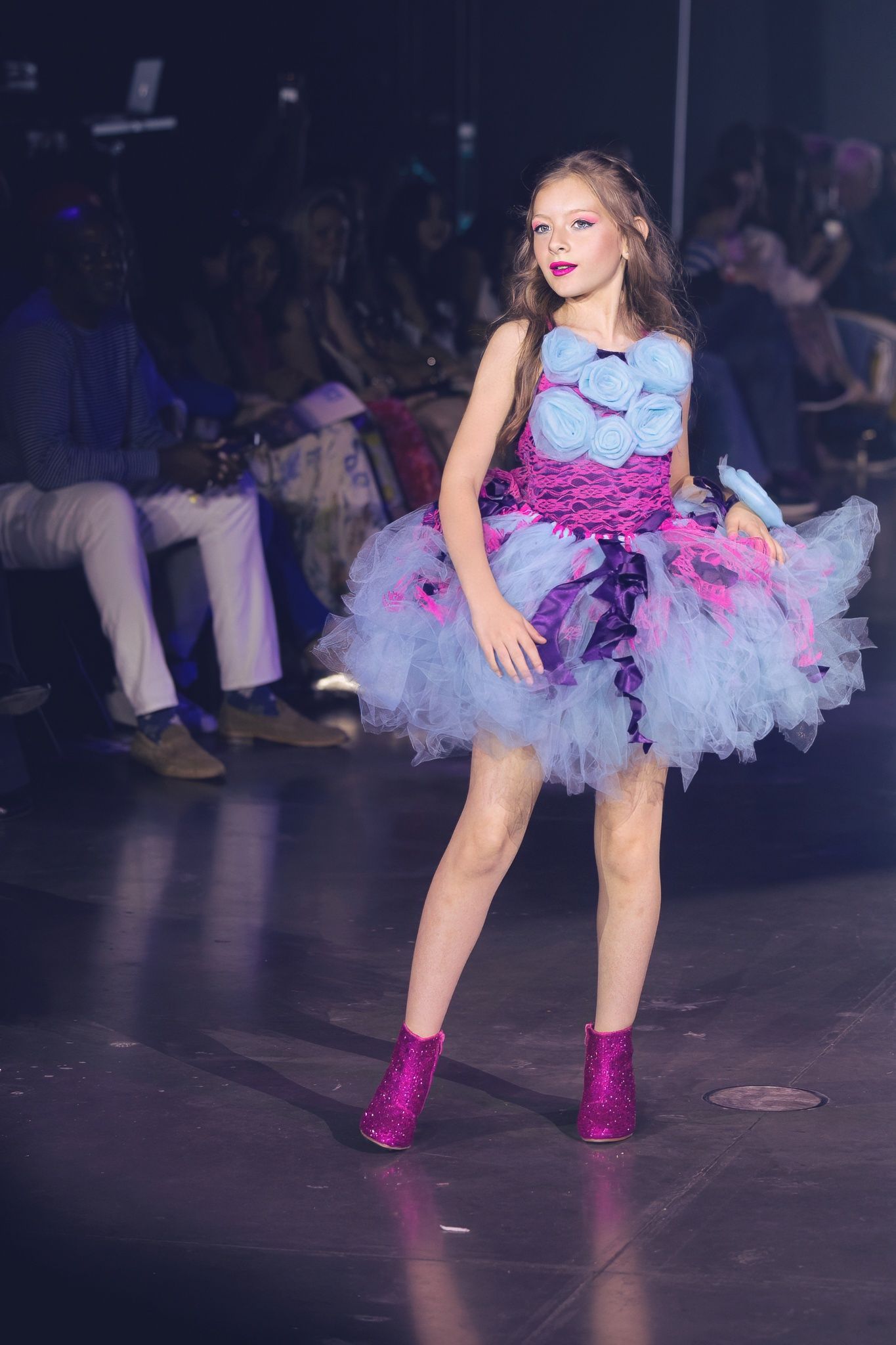 Model in blue and pink tulle dress with sparkly pink boots poses on dark runway during fashion show.