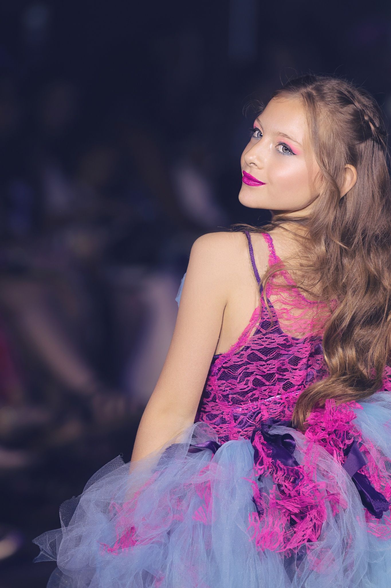 Model wearing a vibrant pink lace and tulle dress with dramatic makeup poses during a fashion show.