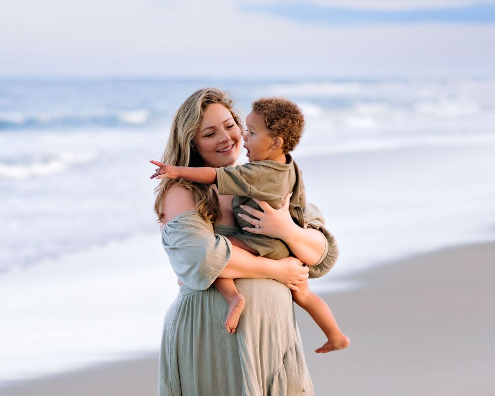 pregnant mom and her toddler son standing on ocean beach interacting with each during outdoor maternity photoshoot