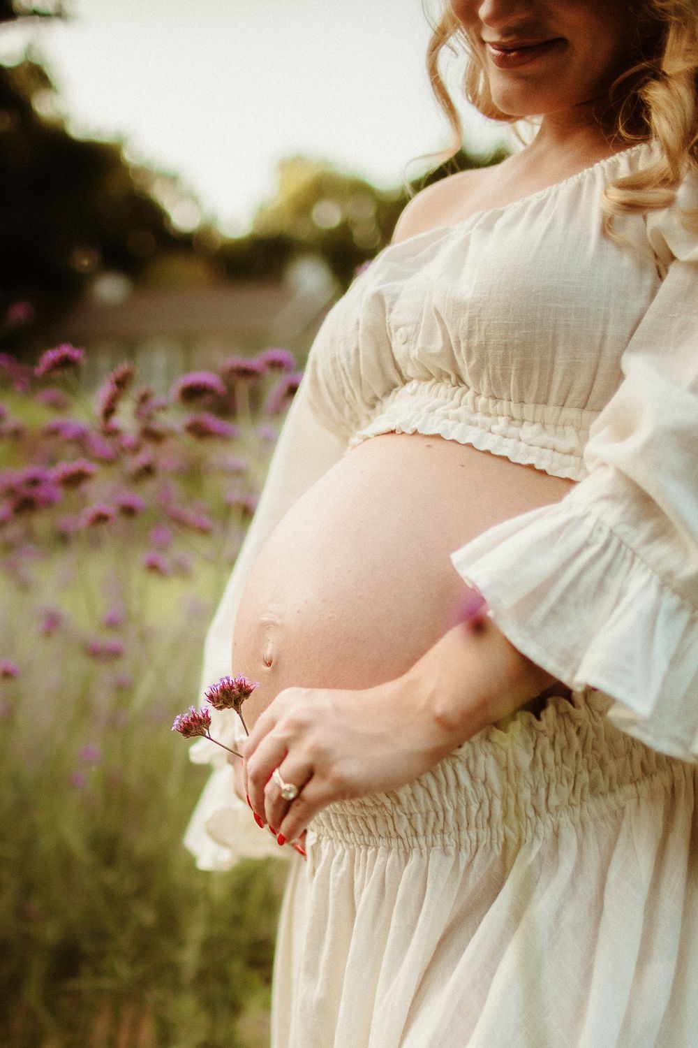 Ethereal maternity photo of pregnant belly in white dress amid purple wildflowers.