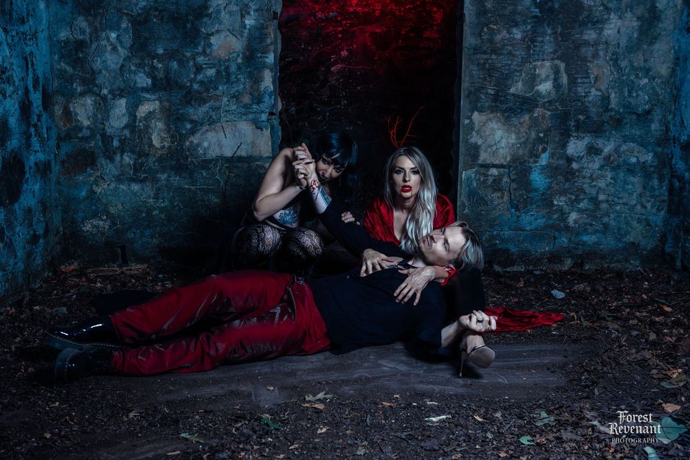 Dark gothic themed photo shoot with two figures in dramatic poses against stone walls.