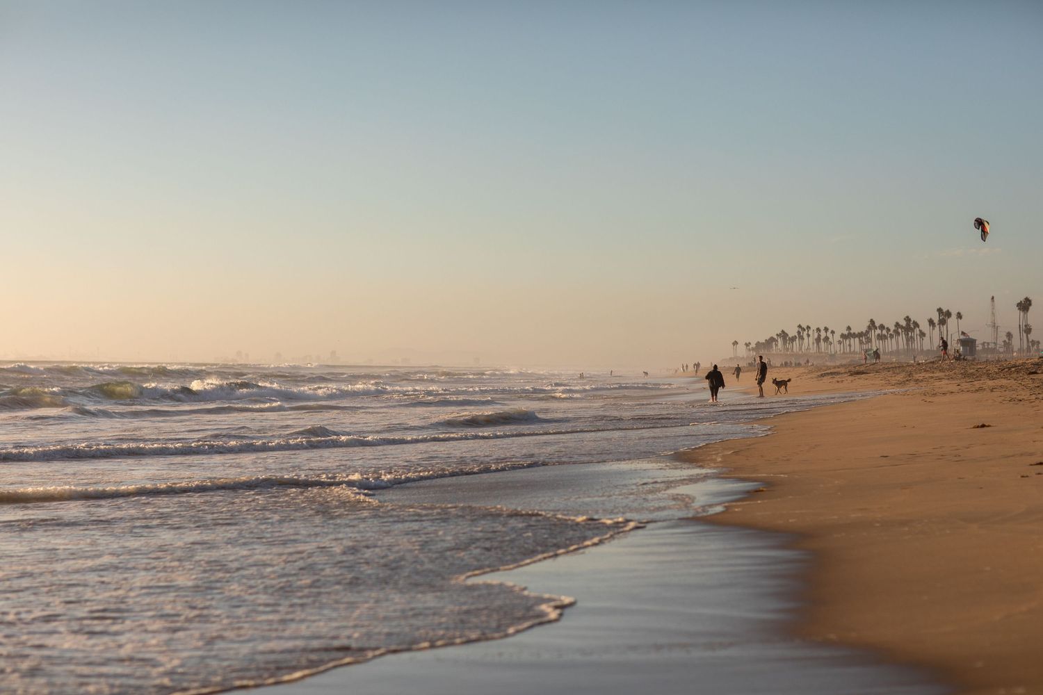 Waves crash on a sandy beach at sunset with palm trees and silhouettes in the distance under a soft sky.