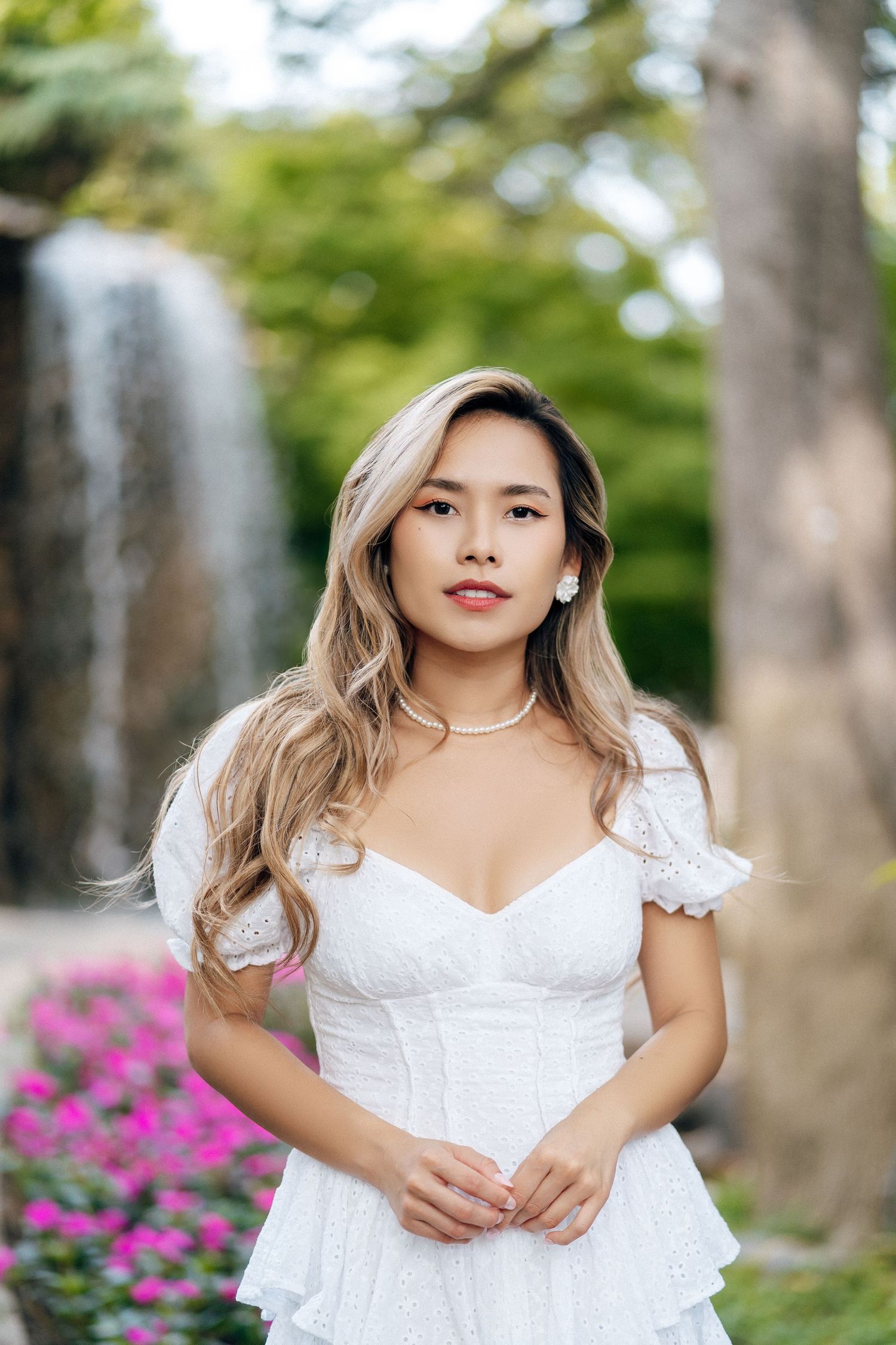 Woman in white lace dress poses outdoors with waterfall and pink flowers in background.
