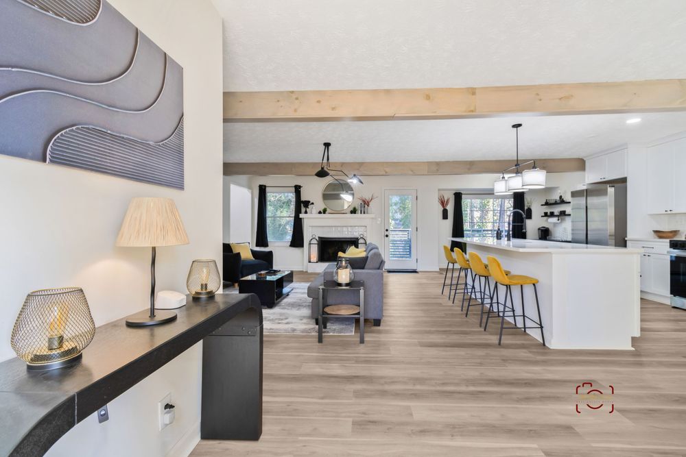 Modern open concept living space with light wood floors, white walls, and a kitchen bar area featuring yellow stools.