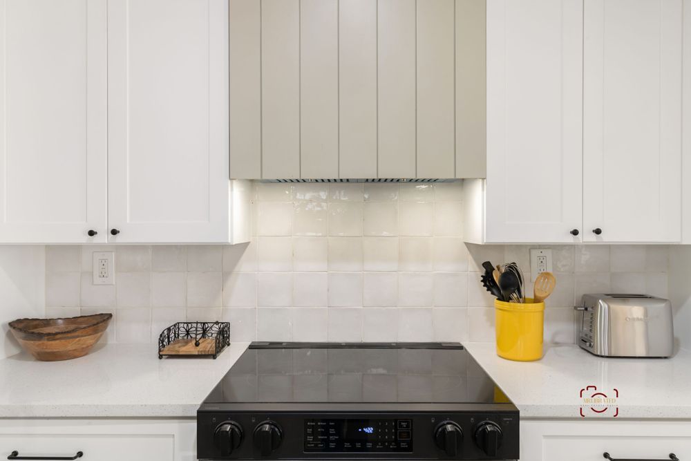 White modern kitchen features stainless steel range hood and black cooktop with white backsplash and cabinets.