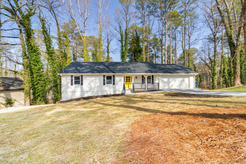 White ranch style home with blue roof sits on grassy landscaped lot surrounded by tall trees.