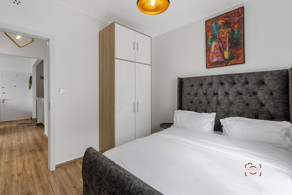 Modern bedroom interior with light wood flooring, built-in white closets, and a dark tufted headboard illuminated by sconces.