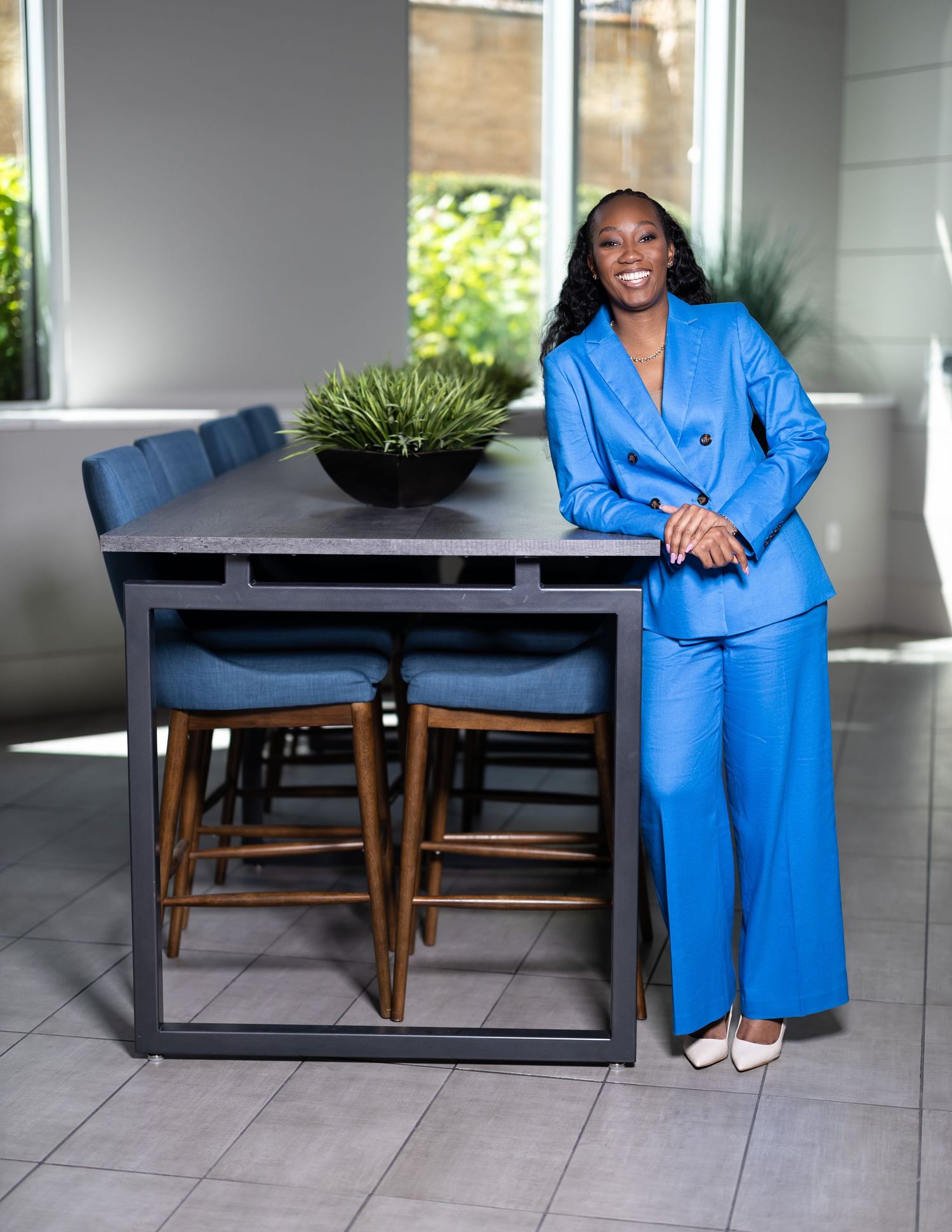 Woman in bright blue pantsuit poses confidently next to modern dining furniture.