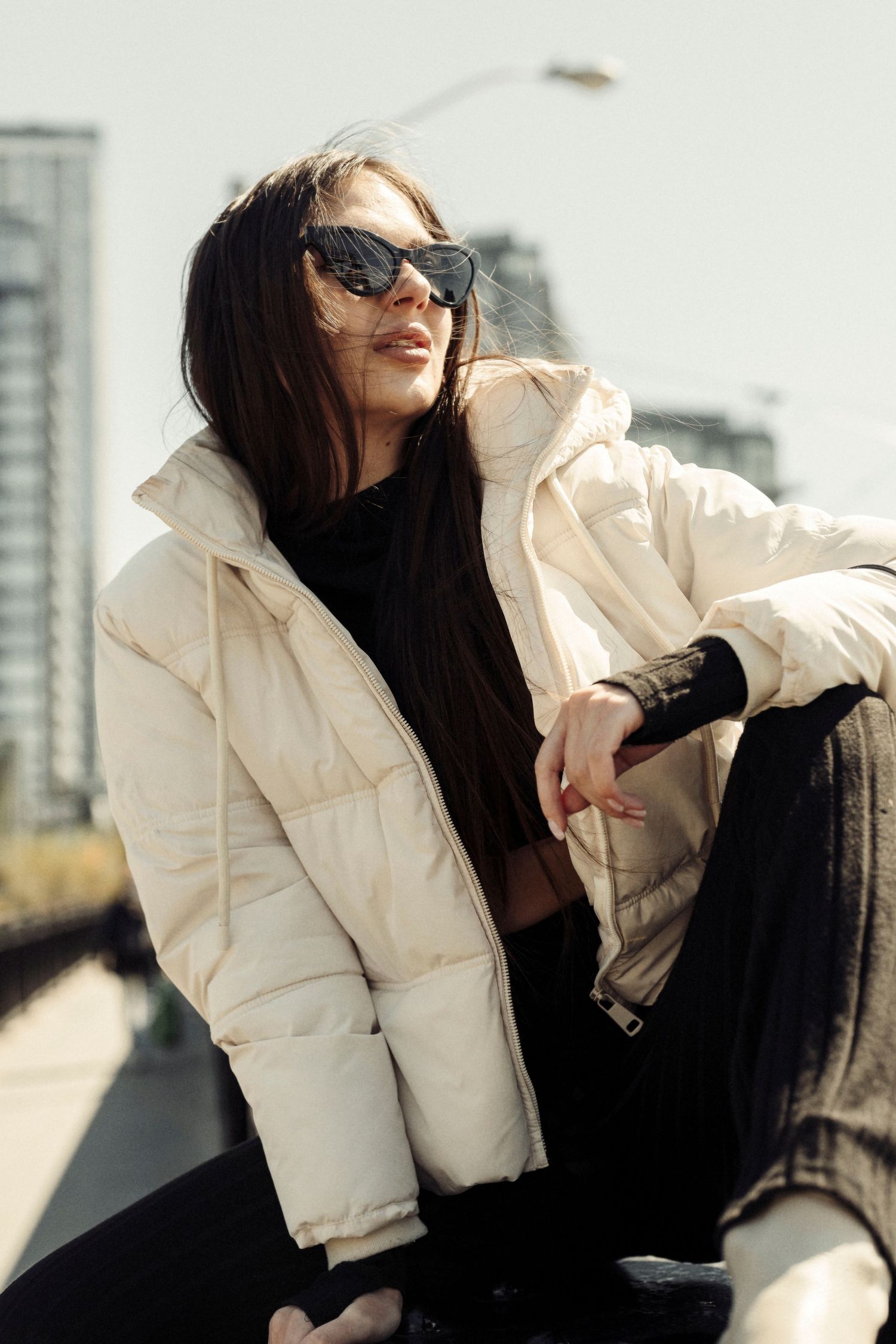 Urban fashion portrait featuring a white puffer jacket and sunglasses against city buildings.