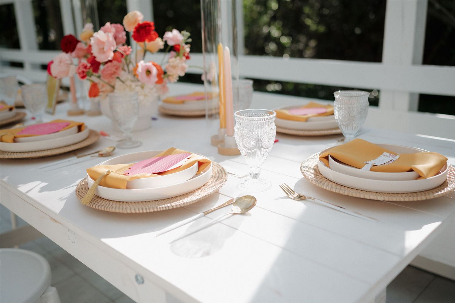 Elegant table setting with pink plates, gold utensils, and coral floral centerpiece in natural sunlight.