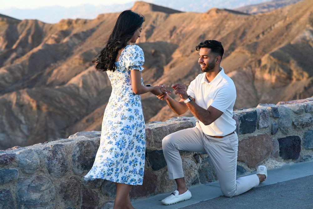 Man proposing to woman on one knee with scenic mountain backdrop at sunset.