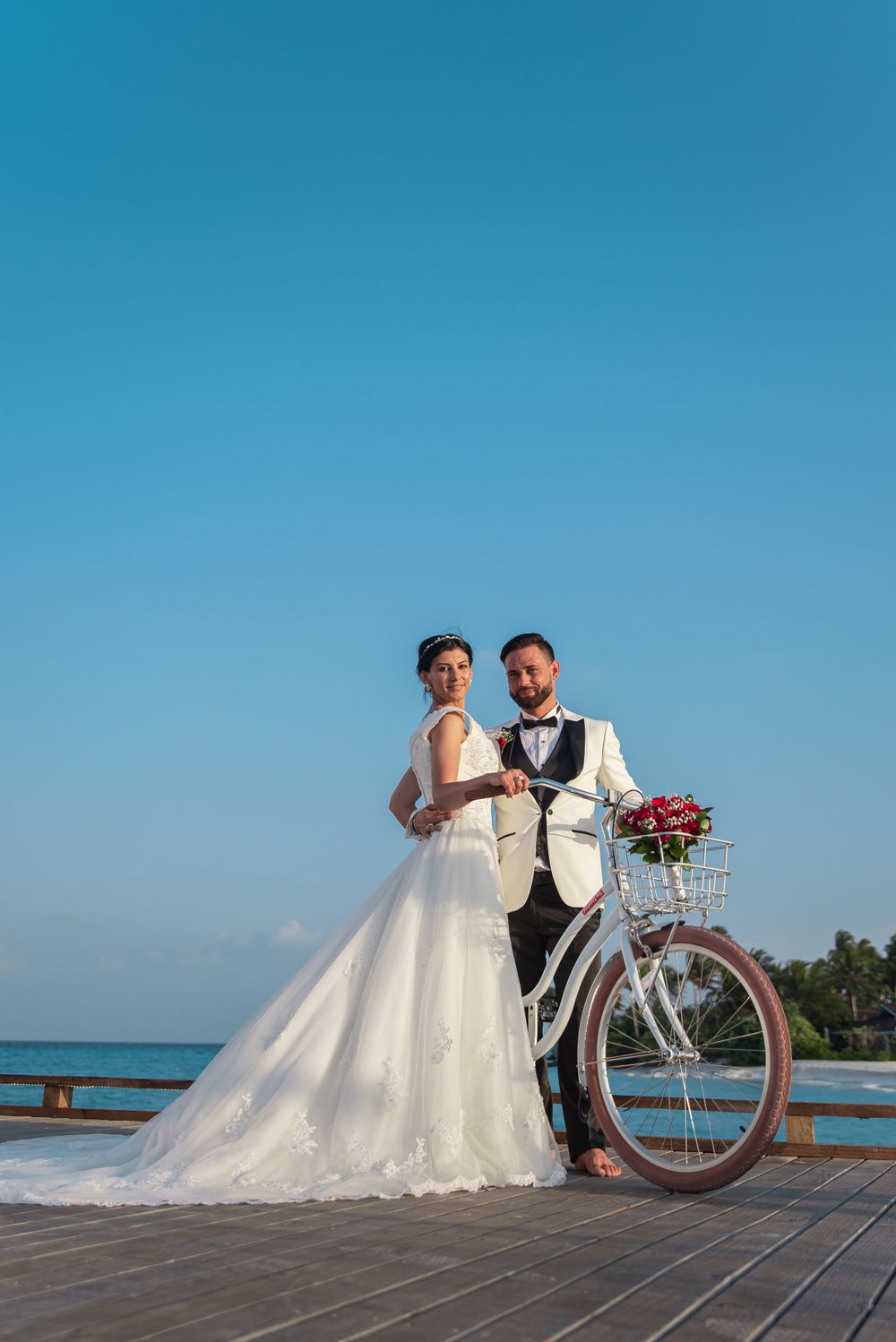 Wedding couple posing with a white bicycle on a seaside deck under blue sky.