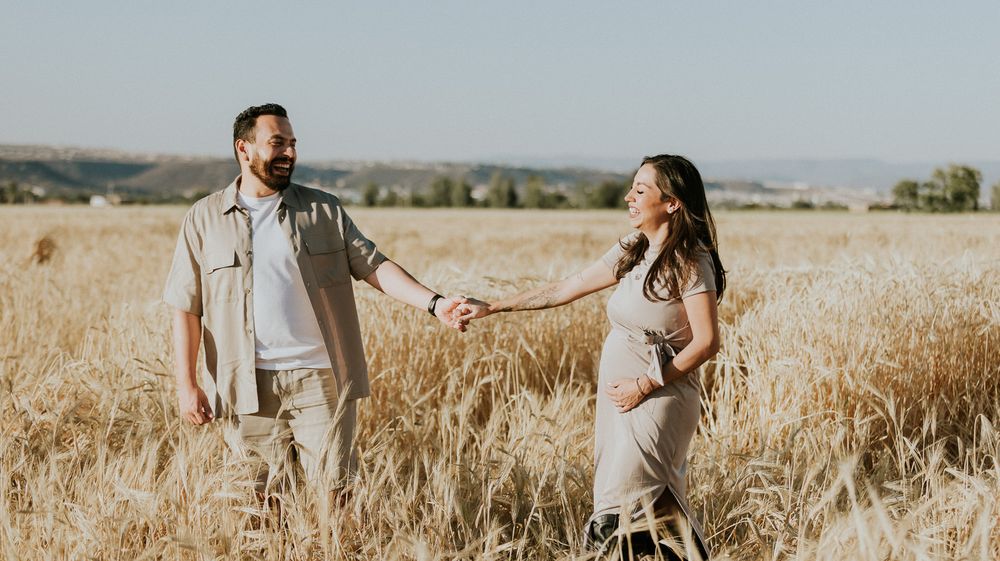 Couple holds hands while walking through tall golden grass with mountains in background.