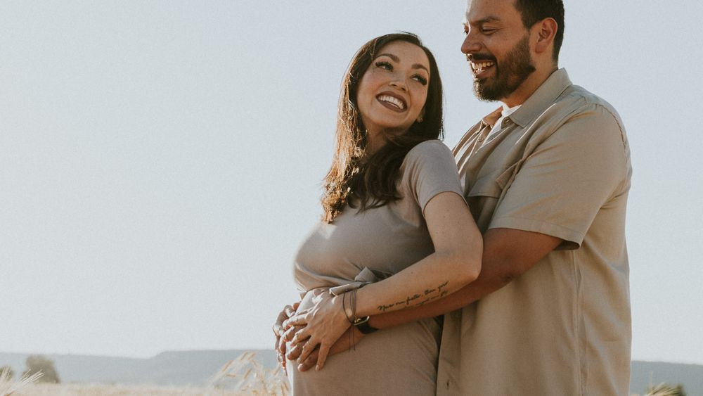 Expecting couple embraces in a golden field during sunset with mountains in background.