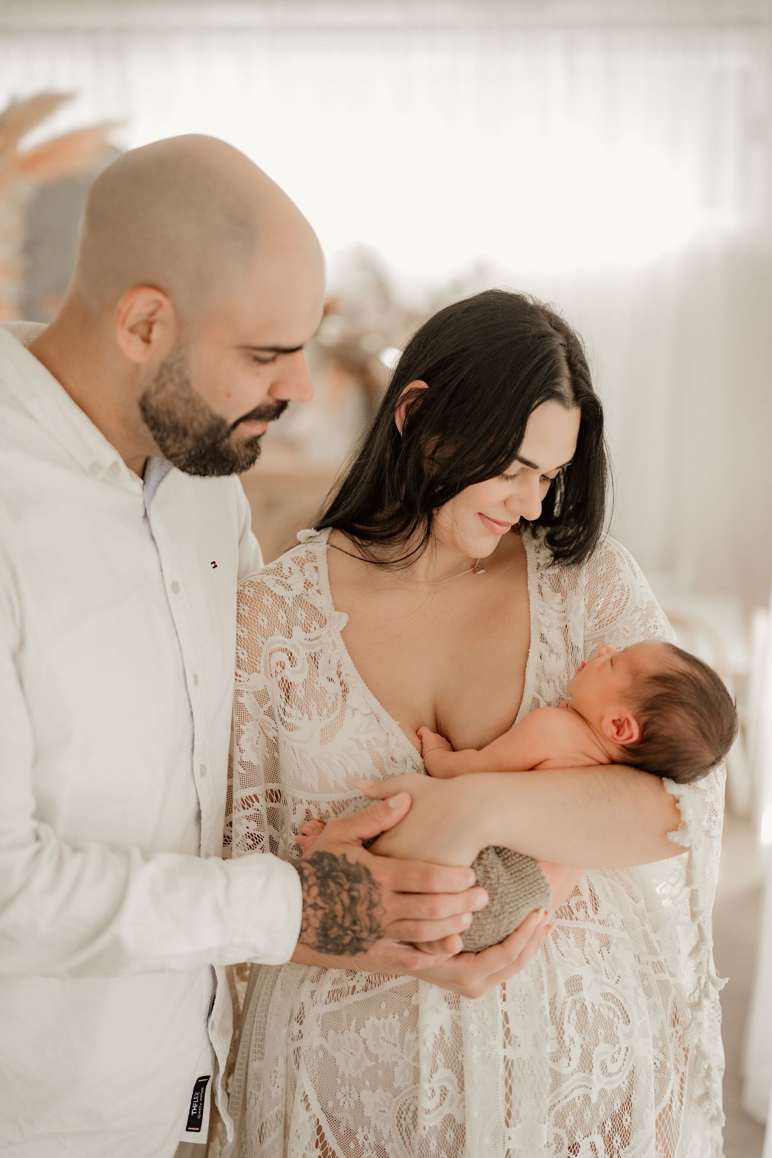 Bald father and dark-haired mother in lace dress lovingly holding newborn baby indoors.