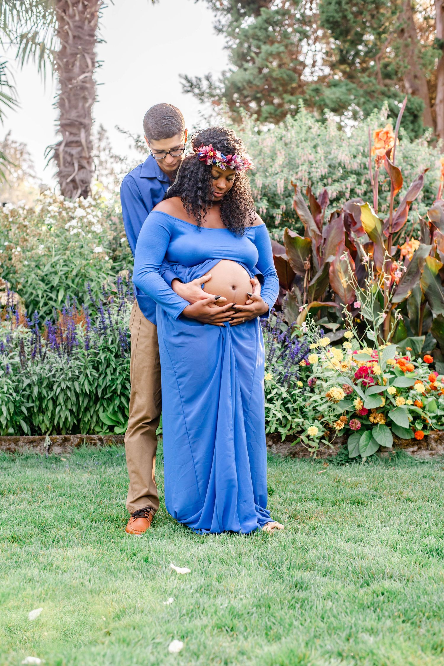 A maternity photoshoot in a garden features a couple in blue and tan embracing among colorful flowers.