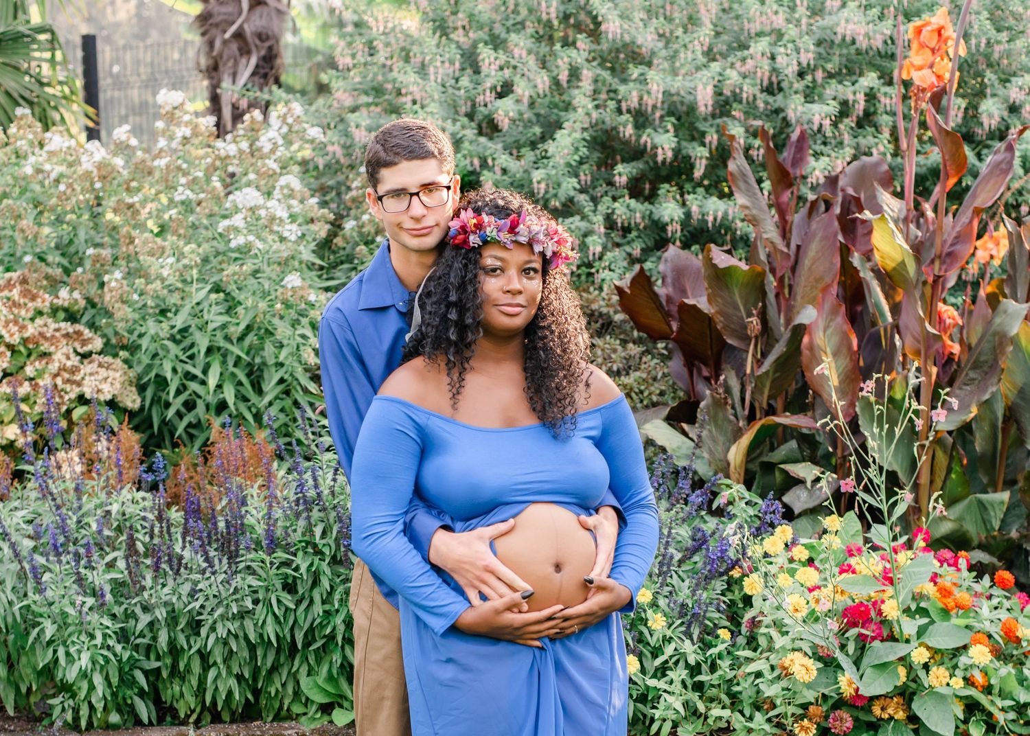 An intimate maternity portrait captures a couple in coordinated blue outfits posing in a lush garden setting.