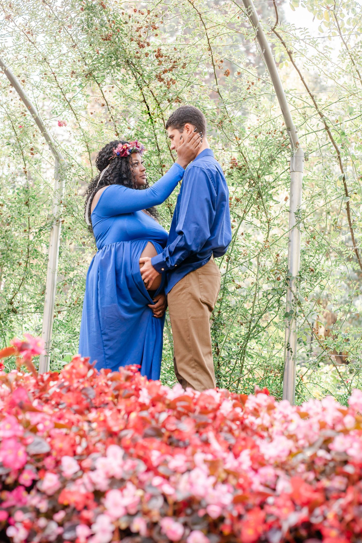 A tender maternity portrait among red flowers features a couple in coordinating blue and tan outfits.