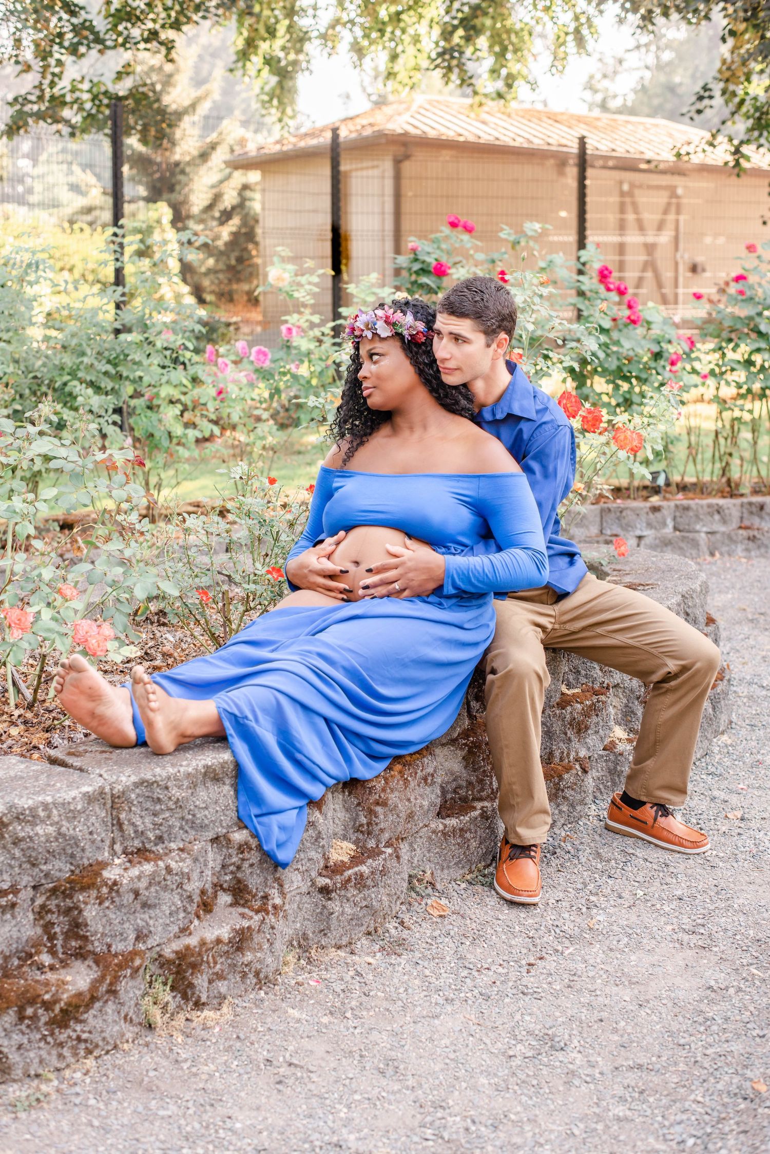 Maternity photo of couple sitting on stone wall in blooming garden with roses.